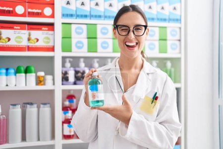 Foto de Mujer morena trabajando en farmacia sosteniendo jarabe para la tos guiñando el ojo mirando a la cámara con expresión sexy, cara alegre y feliz. - Imagen libre de derechos