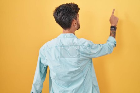Foto de Young hispanic man with tattoos standing over yellow background posing backwards pointing ahead with finger hand - Imagen libre de derechos