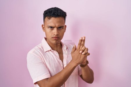 Photo for Young hispanic man standing over pink background holding symbolic gun with hand gesture, playing killing shooting weapons, angry face - Royalty Free Image