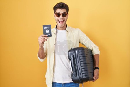 Foto de Joven hispano sosteniendo maleta yendo de vacaciones de verano sosteniendo pasaporte italiano celebrando loco y sorprendido por el éxito con los ojos abiertos gritando emocionado. - Imagen libre de derechos
