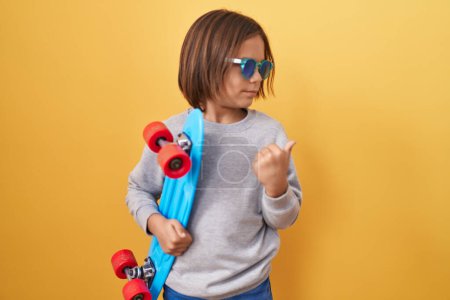 Photo for Little hispanic boy wearing sunglasses holding skate pointing thumb up to the side smiling happy with open mouth - Royalty Free Image