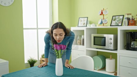 Photo for Young beautiful hispanic woman smelling flowers in a vase at home - Royalty Free Image