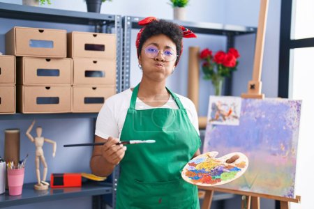 Photo for Young african american woman drawing on canvas at art studio puffing cheeks with funny face. mouth inflated with air, catching air. - Royalty Free Image