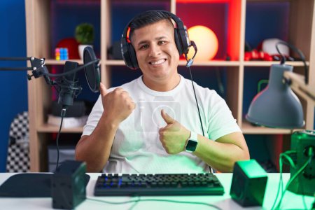 Photo for Young hispanic man playing video games pointing to the back behind with hand and thumbs up, smiling confident - Royalty Free Image
