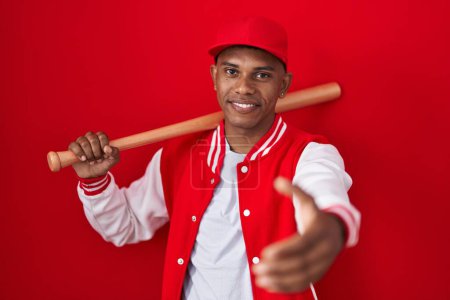 Foto de Joven hispano jugando béisbol sosteniendo bate sonriente amistoso ofreciendo apretón de manos como saludo y bienvenida. negocios exitosos. - Imagen libre de derechos