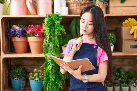 Photo for Young chinese woman florist writing on notebook with doubt expression at flower shop - Royalty Free Image