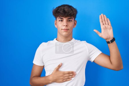 Foto de Adolescente hispano de pie sobre fondo azul jurando con la mano en el pecho y la palma abierta, haciendo un juramento de lealtad promesa - Imagen libre de derechos
