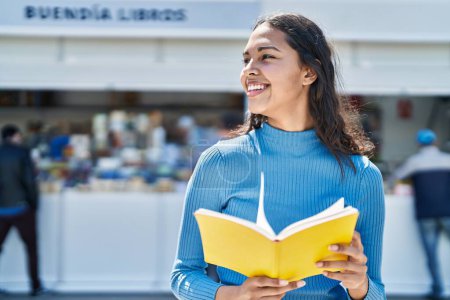 Téléchargez les photos : Jeune femme afro-américaine souriant livre de lecture confiant à la rue - en image libre de droit