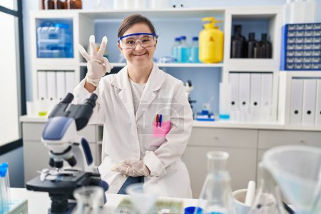 Foto de Chica hispana con síndrome de Down que trabaja en el laboratorio científico sonriendo positiva haciendo signo de ok con la mano y los dedos. expresión exitosa. - Imagen libre de derechos