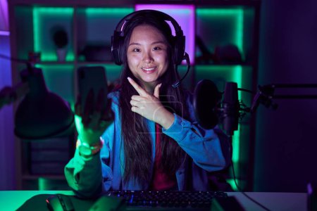 Téléchargez les photos : Jeune femme asiatique jouer à des jeux vidéo avec smartphone gai avec un sourire sur le visage pointant avec la main et le doigt vers le côté avec expression heureuse et naturelle - en image libre de droit