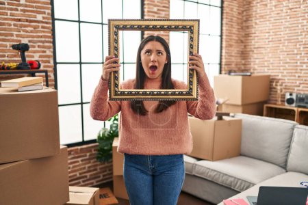 Photo for Young latin woman moving to a new home holding empty frame angry and mad screaming frustrated and furious, shouting with anger looking up. - Royalty Free Image