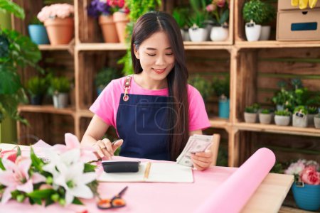 Téléchargez les photos : Jeune femme chinoise fleuriste souriant confiant compter les dollars à la boutique de fleurs - en image libre de droit