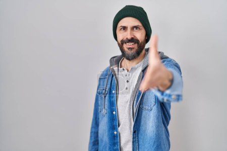 Foto de Joven hombre hispano con tatuajes con gorra de lana sonriente amistoso ofreciendo apretón de manos como saludo y bienvenida. negocios exitosos. - Imagen libre de derechos