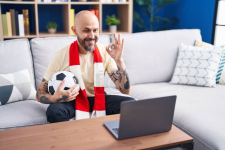 Téléchargez les photos : Homme hispanique avec des tatouages regarder match de football hooligan tenant ballon sur l'ordinateur portable faire ok signe avec les doigts, sourire geste amical excellent symbole - en image libre de droit