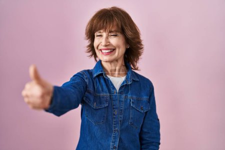 Téléchargez les photos : Femme d'âge moyen debout sur fond rose regardant fier, souriant faire les pouces geste vers le haut sur le côté - en image libre de droit