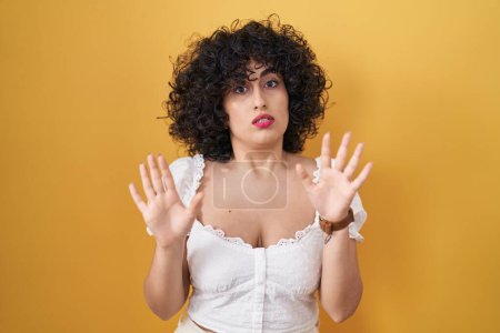 Photo for Young brunette woman with curly hair standing over yellow background moving away hands palms showing refusal and denial with afraid and disgusting expression. stop and forbidden. - Royalty Free Image