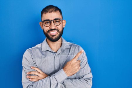 Photo for Middle east man with beard standing over blue background with a big smile on face, pointing with hand and finger to the side looking at the camera. - Royalty Free Image