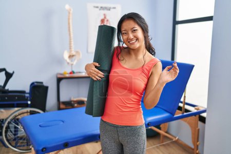 Photo for Young asian woman holding yoga mat at clinic celebrating achievement with happy smile and winner expression with raised hand - Royalty Free Image
