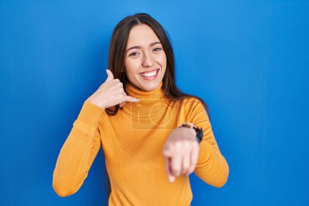 Téléchargez les photos : Jeune femme brune debout sur fond bleu souriant faisant parler au téléphone geste et vous pointant du doigt. Appelle-moi. - en image libre de droit