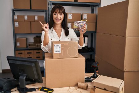 Photo for Young brunette woman working at small business ecommerce holding cart smiling happy pointing with hand and finger to the side - Royalty Free Image