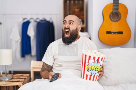 Photo for Young hispanic man with beard and tattoos eating popcorn in the bed angry and mad screaming frustrated and furious, shouting with anger. rage and aggressive concept. - Royalty Free Image