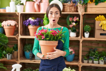 Foto de Mujer hispana joven trabajando en floristería sosteniendo planta asustada y asombrada con la boca abierta para sorpresa, cara de incredulidad - Imagen libre de derechos