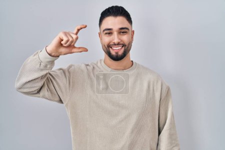 Photo for Young handsome man standing over isolated background smiling and confident gesturing with hand doing small size sign with fingers looking and the camera. measure concept. - Royalty Free Image