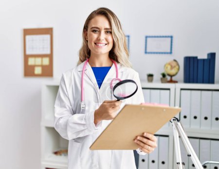 Photo for Young woman wearing doctor uniform holding report using loupe at clinic - Royalty Free Image