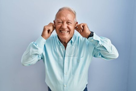Foto de Hombre mayor con el pelo gris de pie sobre el fondo azul cubriendo las orejas con los dedos con expresión molesta por el ruido de la música fuerte. concepto de sordo. - Imagen libre de derechos