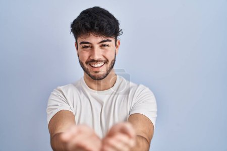 Foto de Hombre hispano con barba de pie sobre fondo blanco sonriendo con las palmas de las manos juntas recibiendo o dando gesto. retención y protección - Imagen libre de derechos