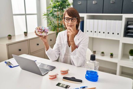Photo for Brunette dentist woman holding sweets serious face thinking about question with hand on chin, thoughtful about confusing idea - Royalty Free Image