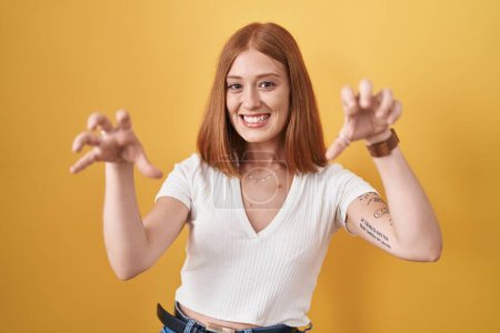 Téléchargez les photos : Jeune rousse femme debout sur fond jaune souriant drôle faisant griffe geste comme chat, expression agressive et sexy - en image libre de droit