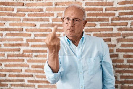 Photo for Senior man with grey hair standing over bricks wall showing middle finger, impolite and rude fuck off expression - Royalty Free Image