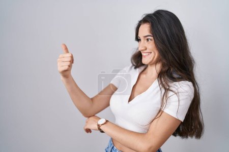 Photo for Young teenager girl standing over white background looking proud, smiling doing thumbs up gesture to the side - Royalty Free Image