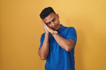 Photo for Young hispanic man standing over yellow background sleeping tired dreaming and posing with hands together while smiling with closed eyes. - Royalty Free Image