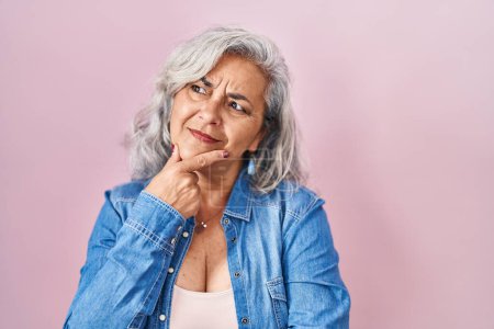 Téléchargez les photos : Middle age woman with grey hair standing over pink background looking confident at the camera smiling with crossed arms and hand raised on chin. thinking positive. - en image libre de droit
