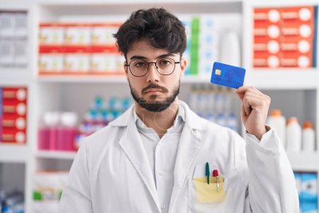 Photo for Hispanic man with beard working at pharmacy drugstore holding credit card thinking attitude and sober expression looking self confident - Royalty Free Image