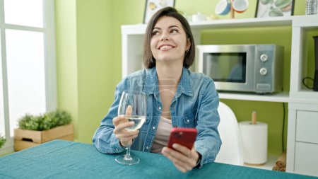 Foto de Mujer joven caucásica usando teléfono inteligente beber vino en casa - Imagen libre de derechos