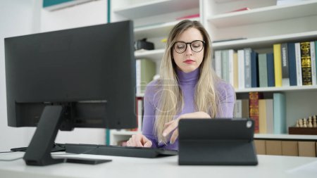 Téléchargez les photos : Jeune étudiante blonde utilisant un ordinateur et un pavé tactile étudiant à la bibliothèque universitaire - en image libre de droit