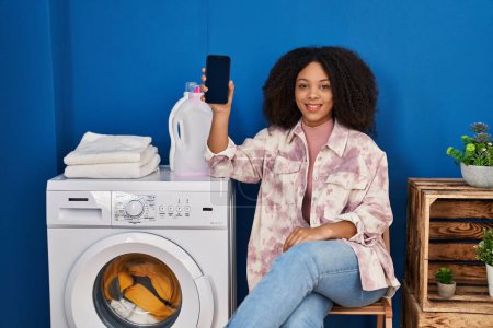 Photo for Young african american woman doing laundry using smartphone looking positive and happy standing and smiling with a confident smile showing teeth - Royalty Free Image