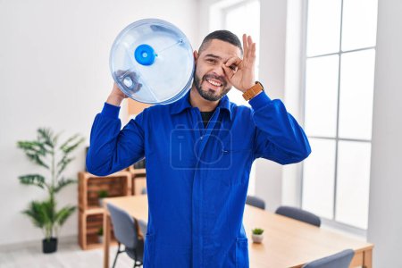 Photo for Hispanic service man holding a gallon bottle of water for delivery smiling happy doing ok sign with hand on eye looking through fingers - Royalty Free Image