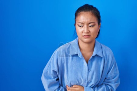 Téléchargez les photos : Asiatique jeune femme debout sur fond bleu avec la main sur le ventre parce que l'indigestion, la maladie douloureuse se sent mal. concept de douleur. - en image libre de droit