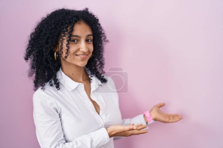 Foto de Mujer hispana con el pelo rizado de pie sobre fondo rosa invitando a entrar sonriendo natural con la mano abierta - Imagen libre de derechos