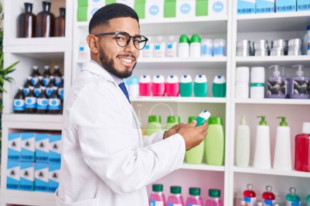 Foto de Joven farmacéutico latino sosteniendo pasta de dientes en estanterías en farmacia - Imagen libre de derechos