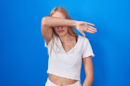 Téléchargez les photos : Jeune femme caucasienne debout sur fond bleu couvrant les yeux avec le bras, l'air grave et triste. concept de non-visibilité, de dissimulation et de rejet - en image libre de droit