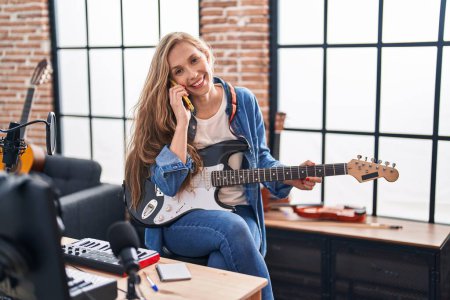 Foto de Joven mujer rubia músico tocando la guitarra eléctrica hablando en el smartphone en el estudio de música - Imagen libre de derechos