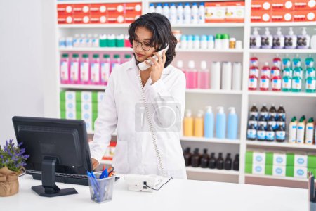 Photo for Young beautiful hispanic woman pharmacist talking on telephone using computer at pharmacy - Royalty Free Image
