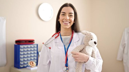 Photo for Young beautiful hispanic woman doctor smiling confident holding teddy bear at clinic - Royalty Free Image
