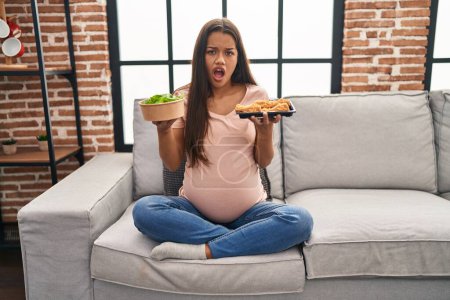 Photo for Young pregnant woman craving for food at home in shock face, looking skeptical and sarcastic, surprised with open mouth - Royalty Free Image