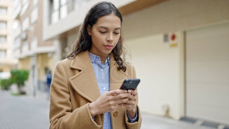 Photo for Young beautiful hispanic woman using smartphone with serious face at street - Royalty Free Image